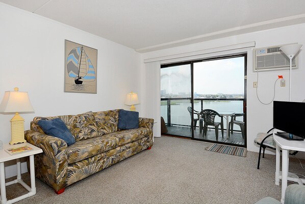 A hotel room with a sofa, two lamps, a TV, and a view of the sea through a glass door. - DAILY ACTIVITIES & LINENS INCLUDED!!! AMAZING BAY & CITY VIEWS!! OUTDOOR POOL! Cozy 1 BR, Right on the Bay! (Ocean City)