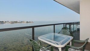 Un balcon avec une table et des chaises en verre, donnant sur une étendue d’eau calme et sur une côte lointaine parsemée de bâtiments.