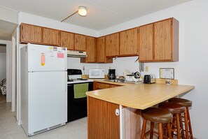 A compact kitchen with wooden cabinets, a white refrigerator, a stove, a microwave, and a coffee maker. There are two stools at the counter.