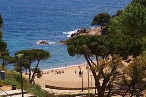 A beach with people sunbathing and walking, surrounded by trees and a rocky coastline.