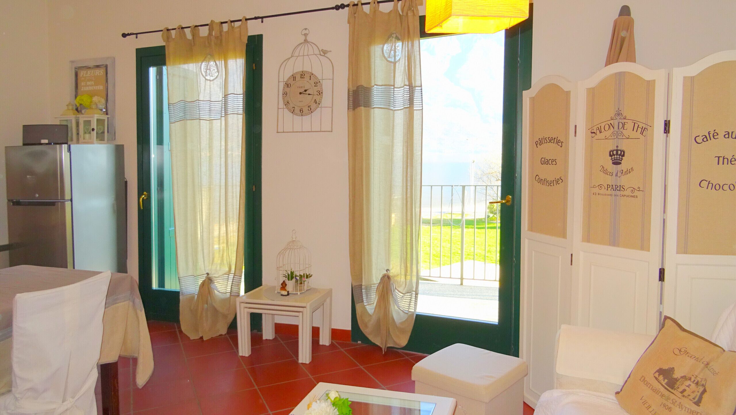 A room with a bed, a small table with a vase, a refrigerator, and a view of the outdoors.