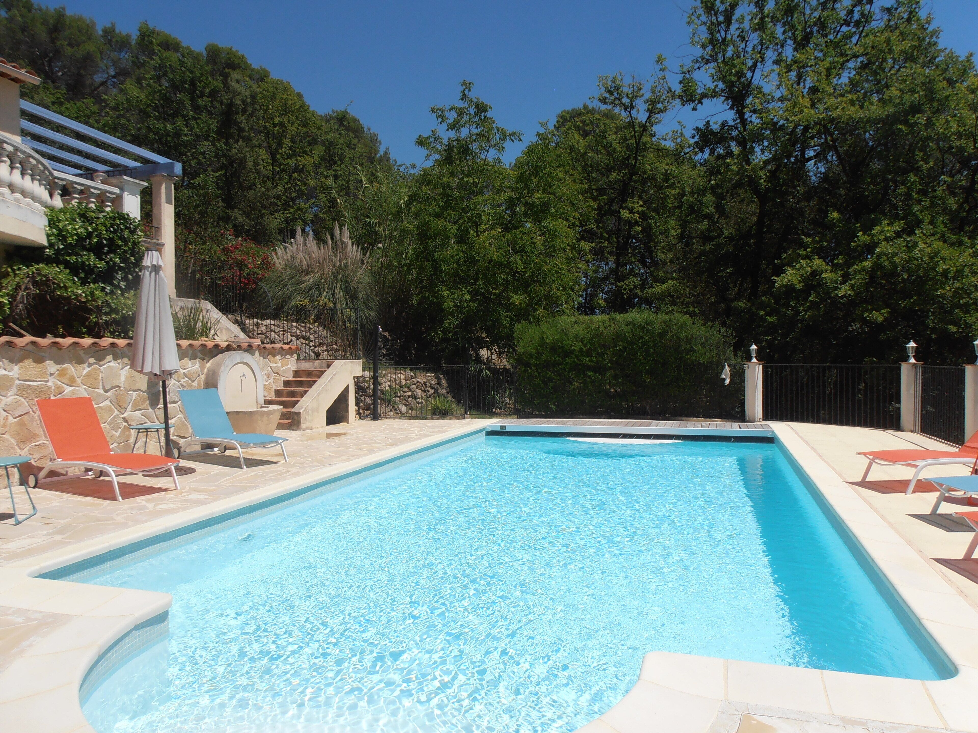 A rectangular swimming pool with clear blue water, surrounded by stone tiles and a stone wall. There are lounge chairs and a pergola with a roof.