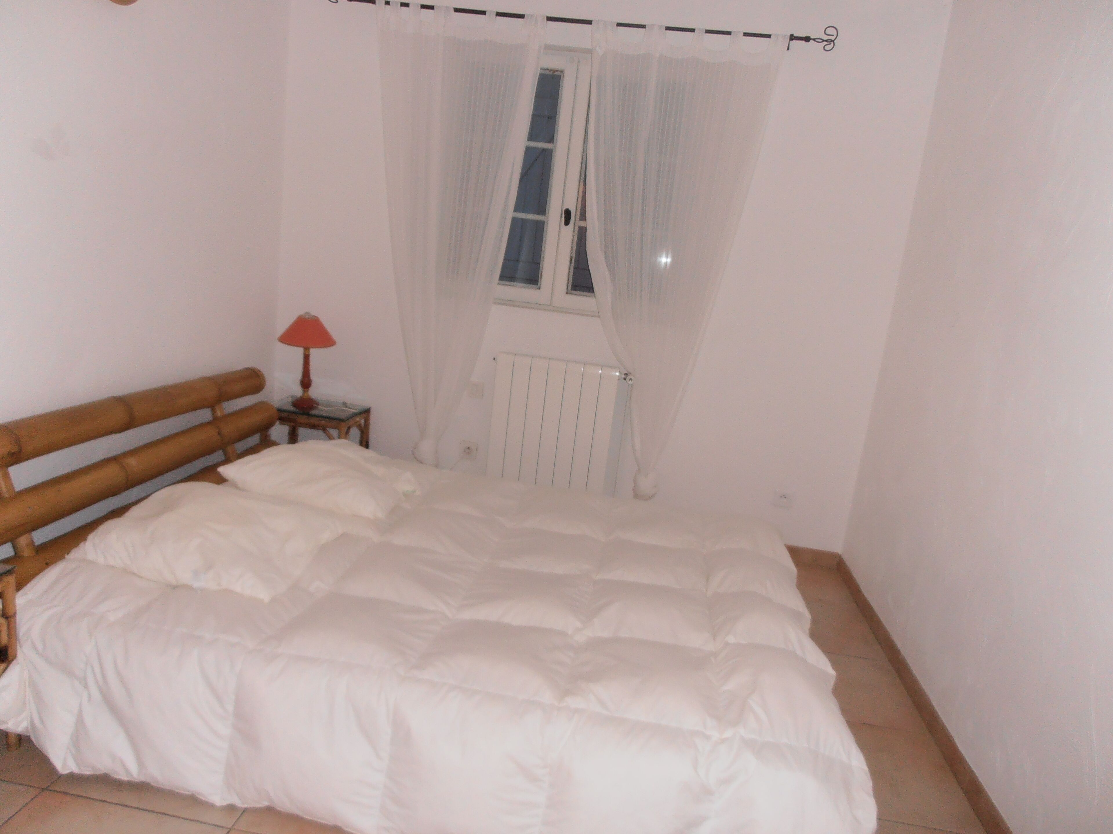 Un dormitorio con una cama de madera, ropa de cama blanca, una mesita de noche pequeña con una lámpara, una ventana con cortinas de encaje y un radiador.