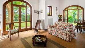 A spacious living room with a floral sofa, wooden furniture, and large arched windows offering a view of greenery.