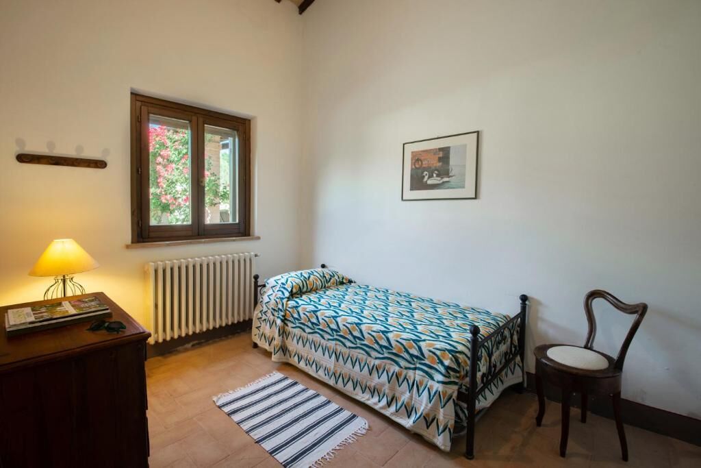 A small bedroom with a single bed, a chair, a table with a lamp, a radiator, a window with a view of flowers, and a framed picture on the wall.