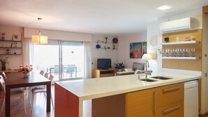 A modern kitchen with a central island, a dining area with chairs, a television, and a window with curtains. - FANTASTIC BEACH FRONT APARTMENT IN SALOU 068 BAHIA (Salou)