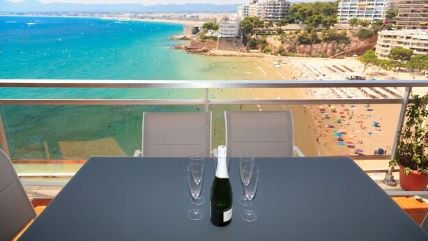 A balcony with a table, two wine glasses, and a bottle of champagne. The view includes a beach, ocean, and buildings. - FANTASTIC BEACH FRONT APARTMENT IN SALOU 068 BAHIA (Salou)
