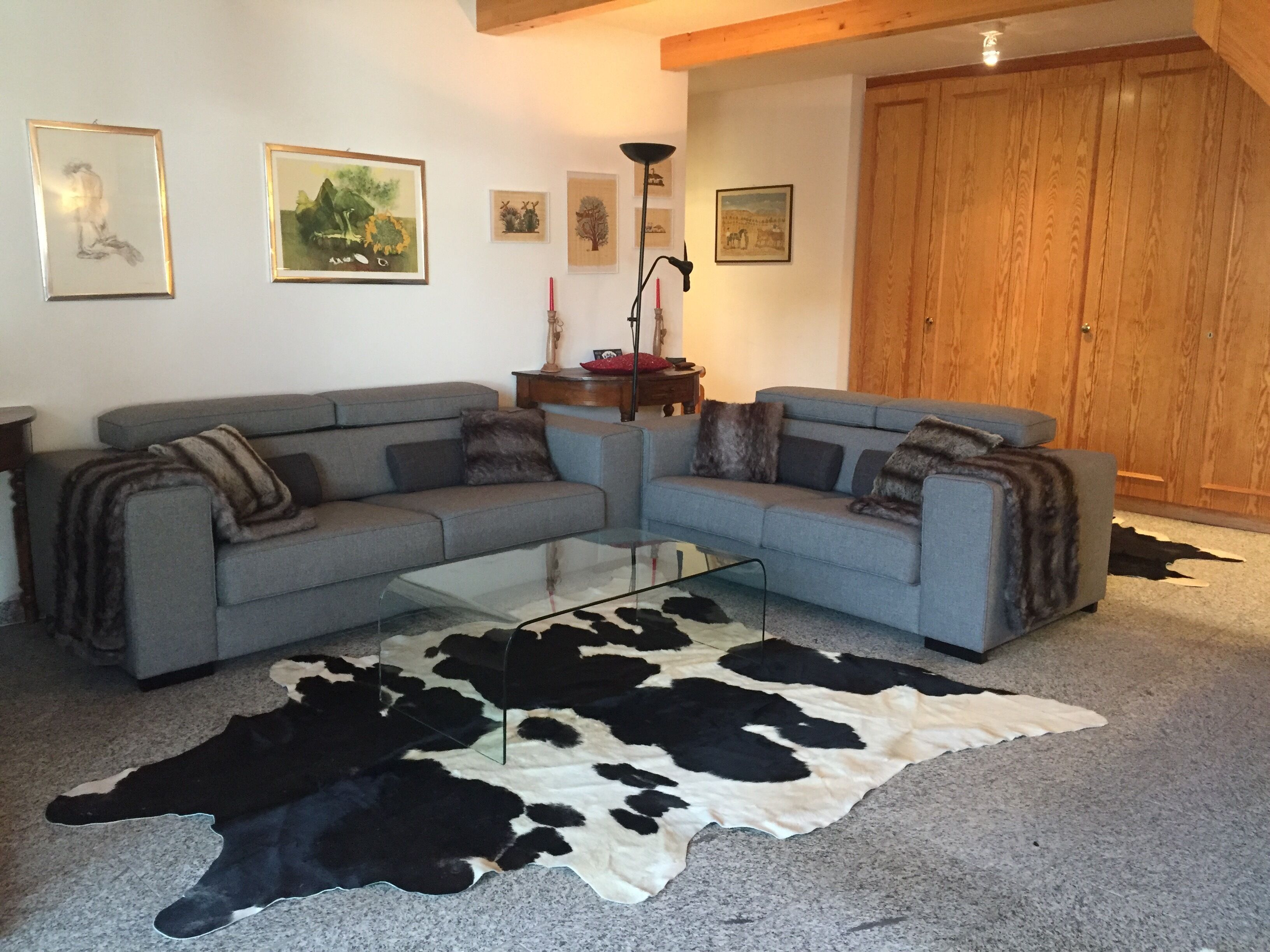 Un salon avec un canapé d’angle gris, une table basse en verre et un tapis en peau de vache noir et blanc. Des tableaux encadrés ornent le mur, et une porte en bois est visible au fond.