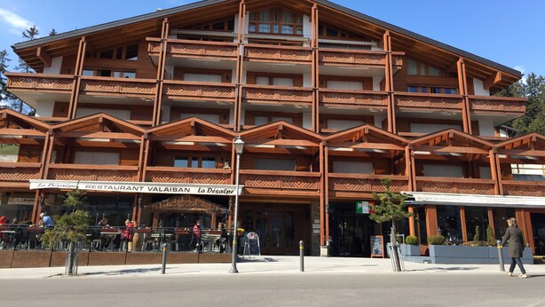 Un edificio de madera de varios pisos con balcones, un restaurante llamado "La Desalpe" en la planta baja y mesas al aire libre.