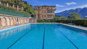 Una piscina con agua azul cristalina, rodeada por un muro de piedra y una fila de sillas de descanso. Al fondo, un edificio de varios pisos con balcones y vistas a la montaña.