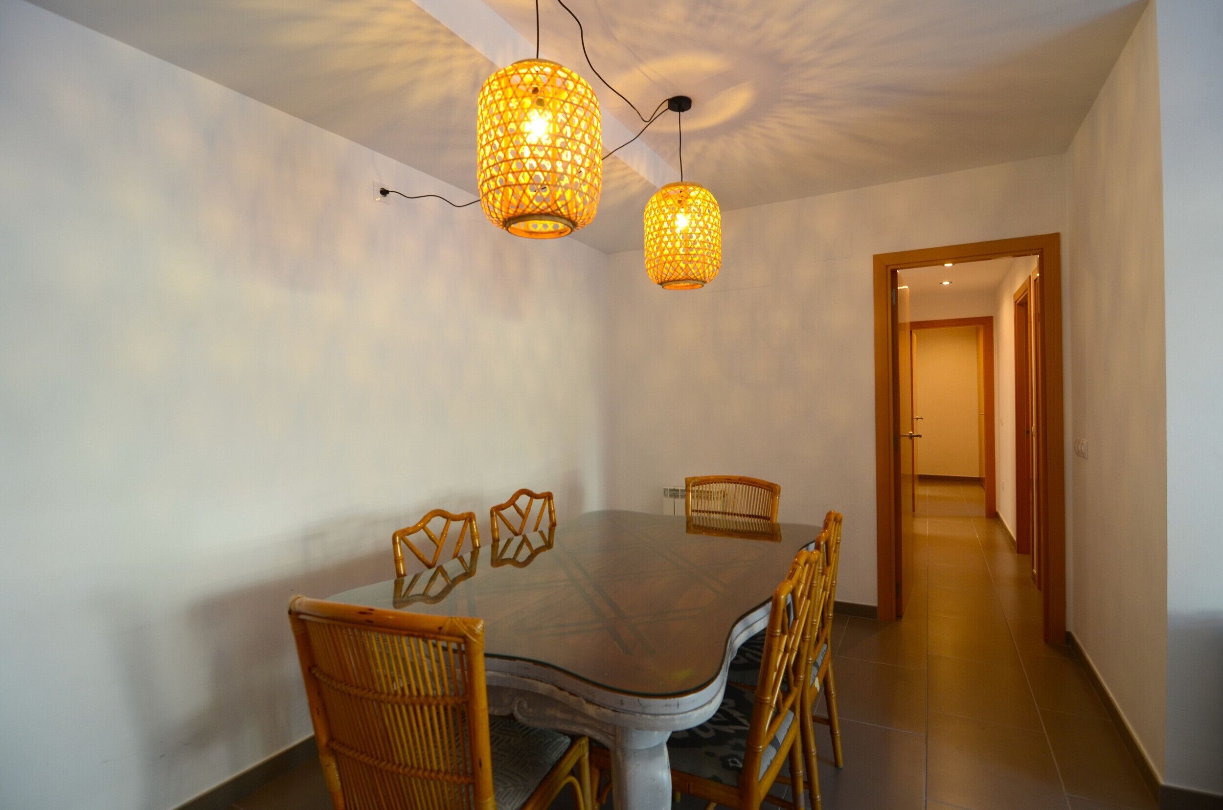 Un comedor con mesa de vidrio, sillas de mimbre y dos lámparas colgantes de estilo farol.