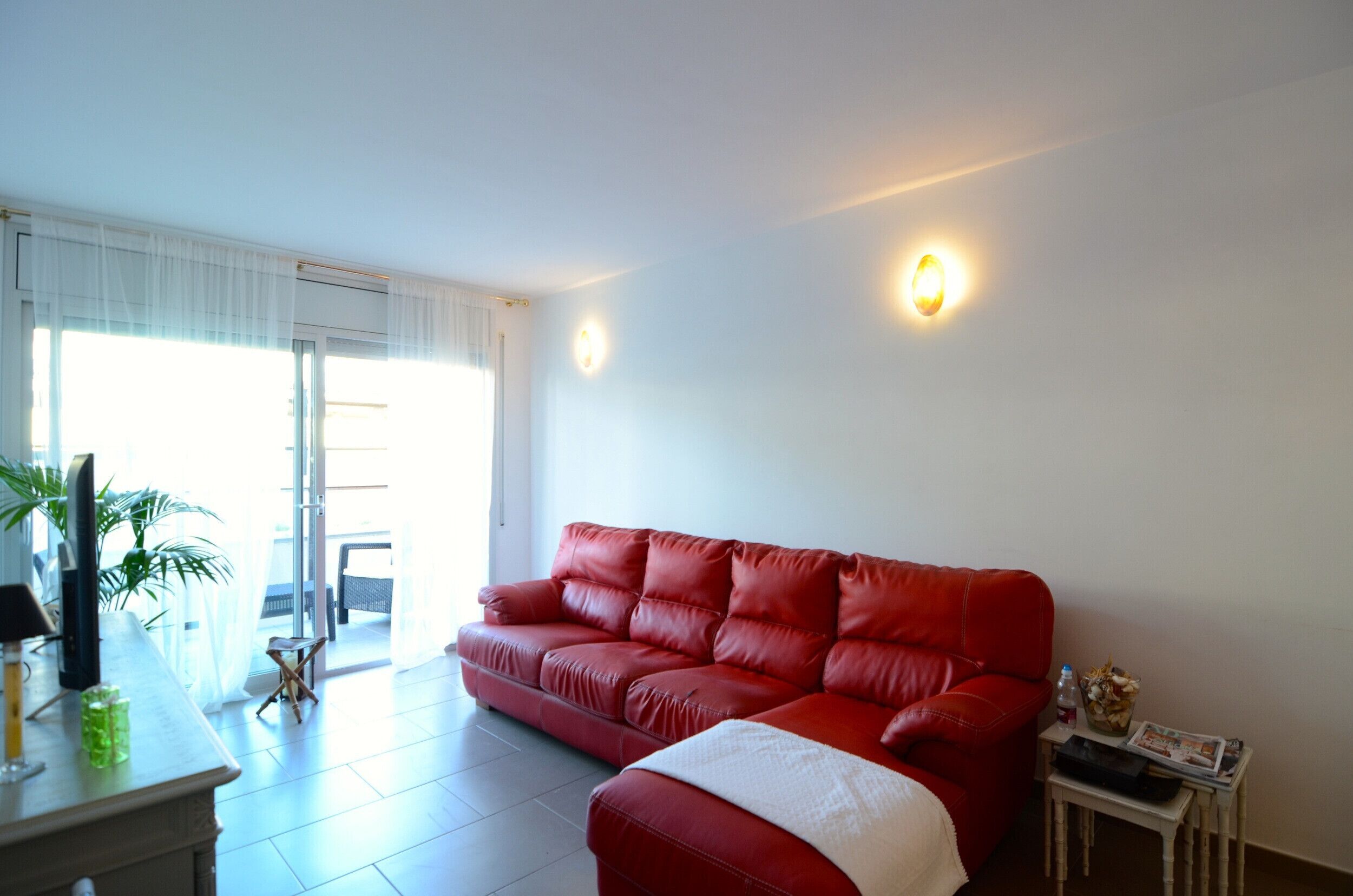 A red sofa in a room with a white wall, a small table with a lamp, and a potted plant.