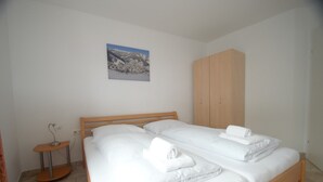 Een tweepersoonsbed met witte lakens en handdoeken, een houten kledingkast, een klein tafeltje met een lamp, en een lijst met een afbeelding aan de muur.