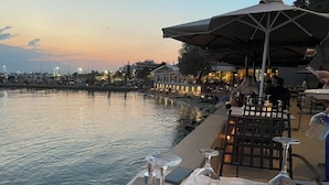 A waterfront dining table set with glasses, a candle, and a menu, overlooking a calm body of water with city lights in the background.