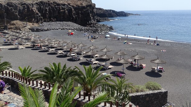 A beach with numerous sun umbrellas and lounge chairs, rocky cliffs, and a clear view of the ocean.