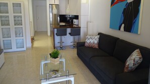 A modern living room with a black sofa, a glass coffee table, and a kitchen in the background.