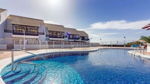 A swimming pool with a clear blue water surface, surrounded by a white fence and a building with a tiled roof.