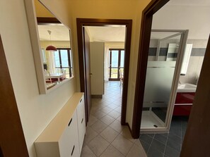 A corridor leading to a bathroom with a shower, a dining area with a table and chairs, and a view of the outdoors.