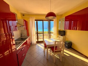 A modern kitchen with red cabinets, a dining area with a table and chairs, and a view of the sea through large glass doors.
