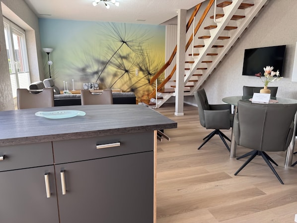 Una cocina moderna con una isla integrada, comedor con sillas, una escalera y un televisor de pantalla plana montado en la pared.