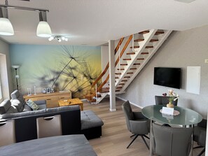 En moderne stue med sofa, skammel, sofabord i glas og et spiseområde med stole. En trappe med trægelænder fører op til en etage.