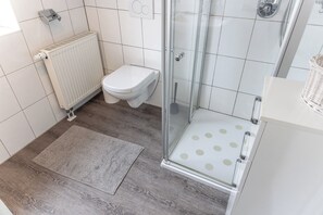A bathroom with a toilet, a shower with a glass door, a radiator, and a patterned rug. - House "dyke Rose" Quiet location, Wi-Fi, 300 meters to the beach, top is directing (Harlesiel)