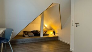 Een slaapkamer met een schuine kap, een bed, een stoel en een deur.