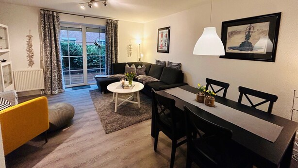 Een moderne woonkamer met een eethoek, een bank, een salontafel en een eettafel met stoelen. Er is een groot raam met gordijnen, een radiator en een plafondventilator.
