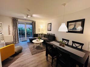 Ein modernes Wohnzimmer mit Essbereich, einer Couch, einem Couchtisch und einem Esstisch mit Stühlen. Es gibt ein großes Fenster mit Vorhängen, eine Heizung und einen Deckenventilator.