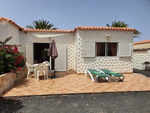 Une petite maison avec un patio carrelé, deux fauteuils de jardin, une table et une porte.