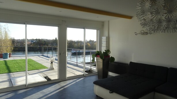 Una sala de estar moderna con un amplio ventanal corredera que da a un balcón con vistas a un río y al paisaje urbano.