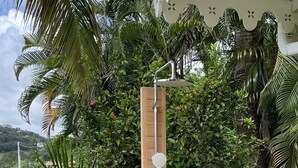 Estructura vertical de madera con cabezal de ducha y manguera, rodeada de vegetación exuberante y una terraza de madera.