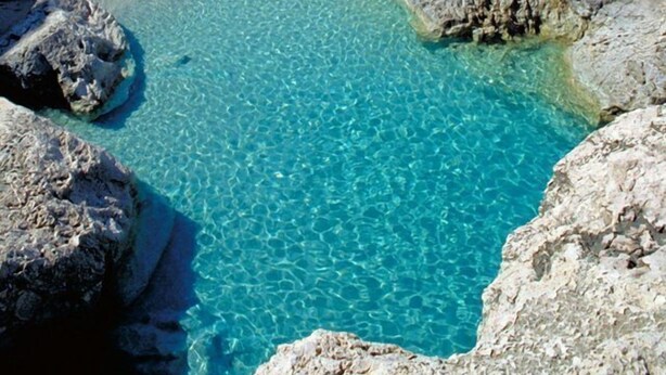 Ein Küstenstrand mit klarem türkisblauem Wasser, Felsformationen und einem gebirgigen Hintergrund.