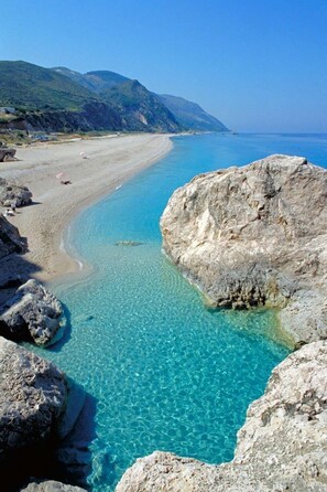Una playa costera con aguas turquesas cristalinas, formaciones rocosas y montañas al fondo.