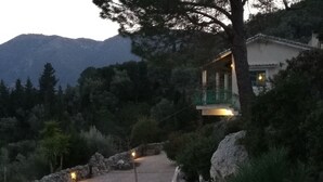 Ein Steinweg führt zu einem Gebäude mit Balkon, umgeben von Bäumen und Bergen im Hintergrund.