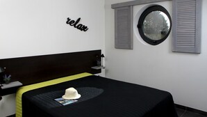 Un lit bien fait, recouvert d’une couvre-lit noire et jaune, un chapeau blanc posé sur le lit, et un grand miroir rond fixé au mur, au-dessus d’une fenêtre à volets.