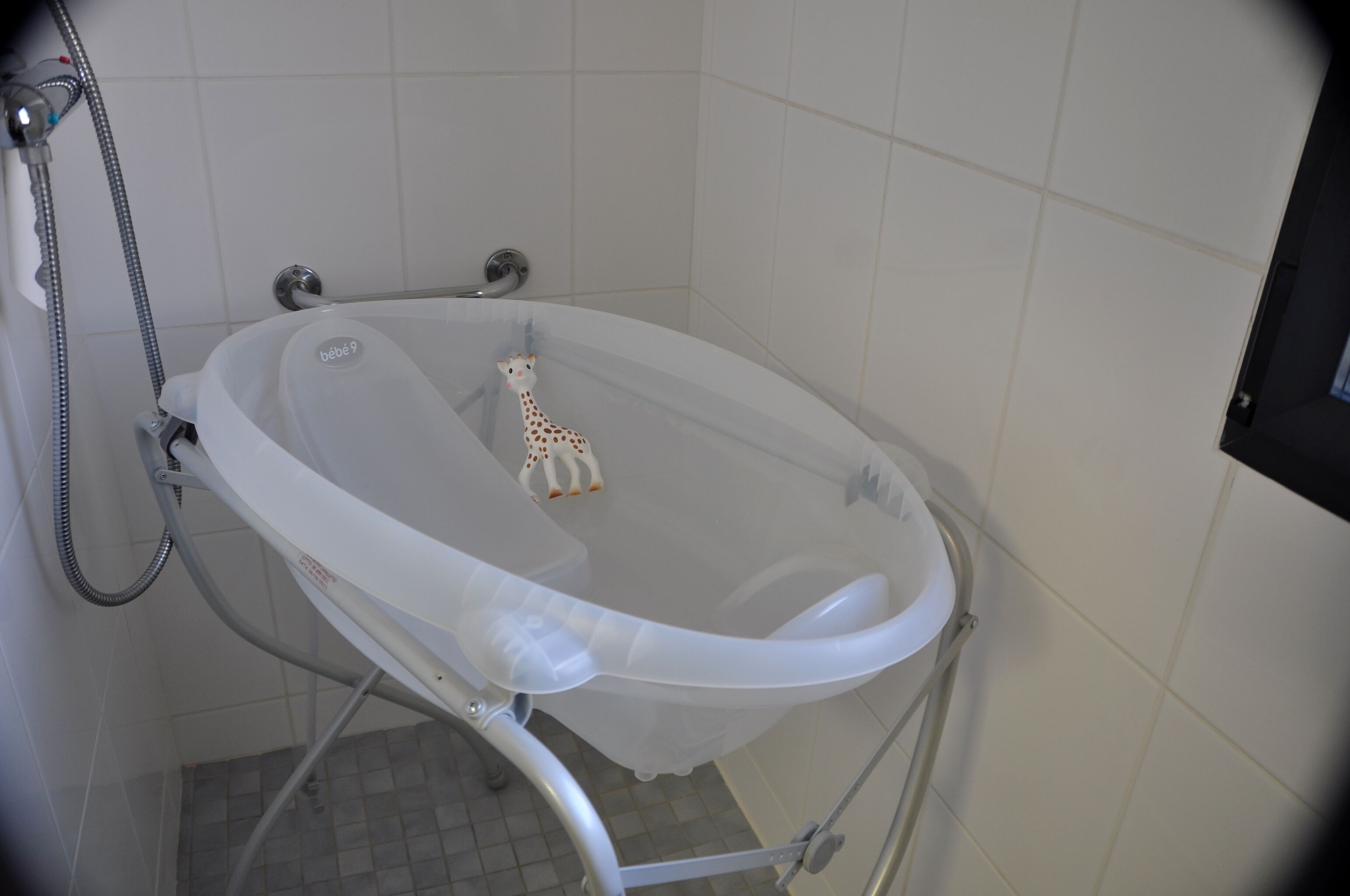 A baby bathtub with a giraffe toy inside, a showerhead, and a tiled floor.