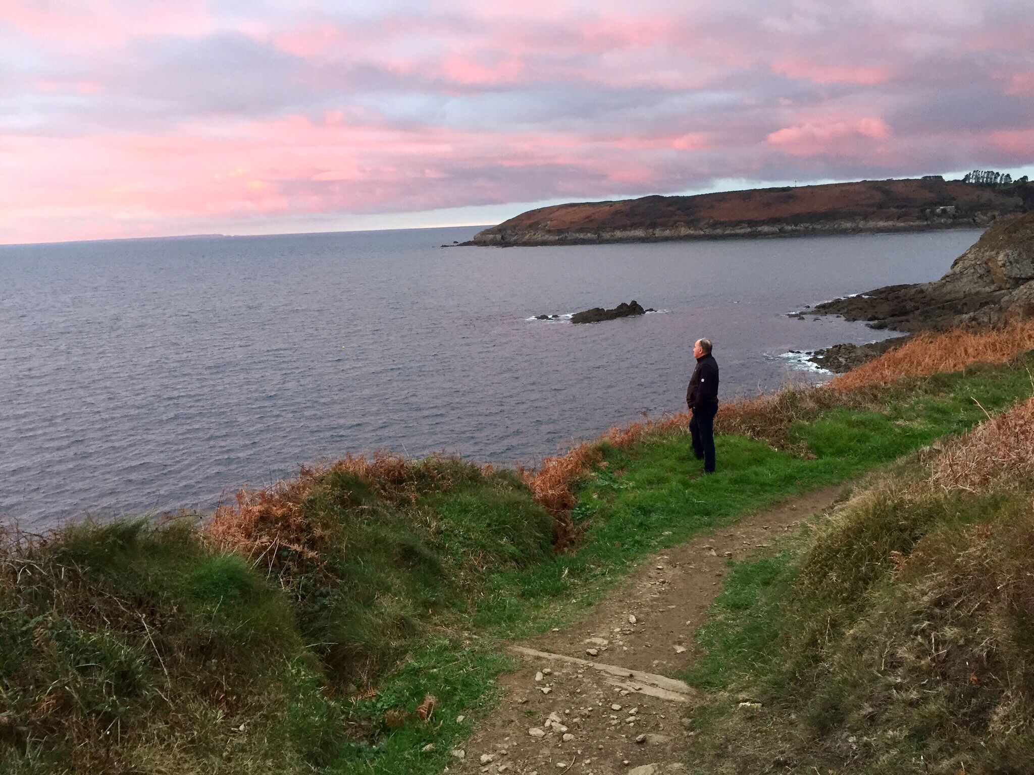 Un camino costero con una persona al borde, mirando al mar y colinas distantes.