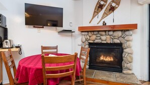 Ein gemütliches Zimmer mit Kamin aus Stein, einem Flachbildfernseher und einem Essbereich mit Holzsesseln und einer roten Tischdecke.