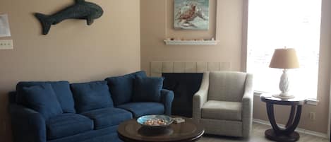 Una sala de estar con un sofá azul, un sillón estampado, una mesita baja con un cuenco, un colgador de pared con forma de pez decorativo, un cuadro sobre la chimenea y una lámpara en una mesita auxiliar.