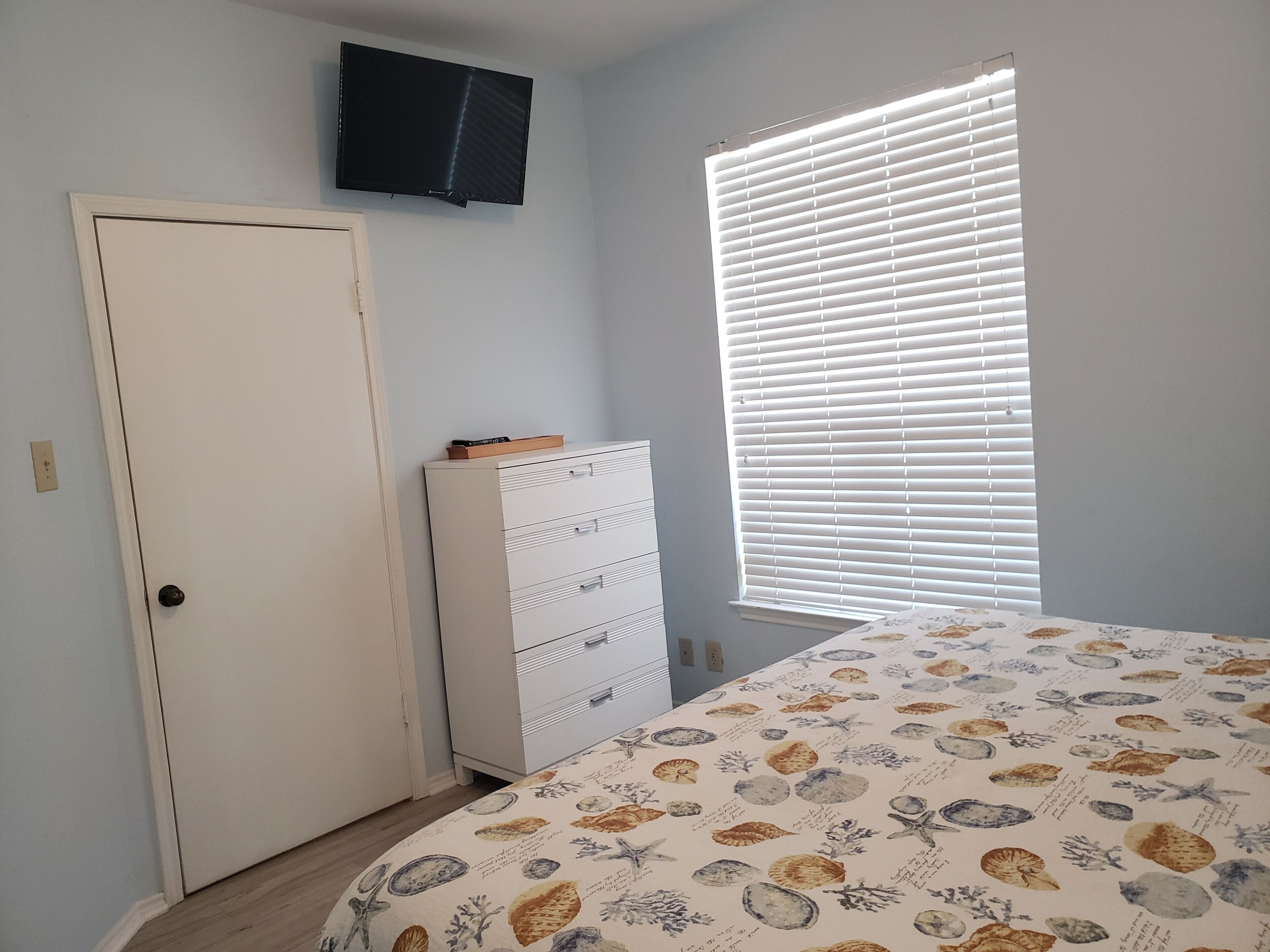 A bedroom with a bed, a dresser, a door, and a window with blinds.