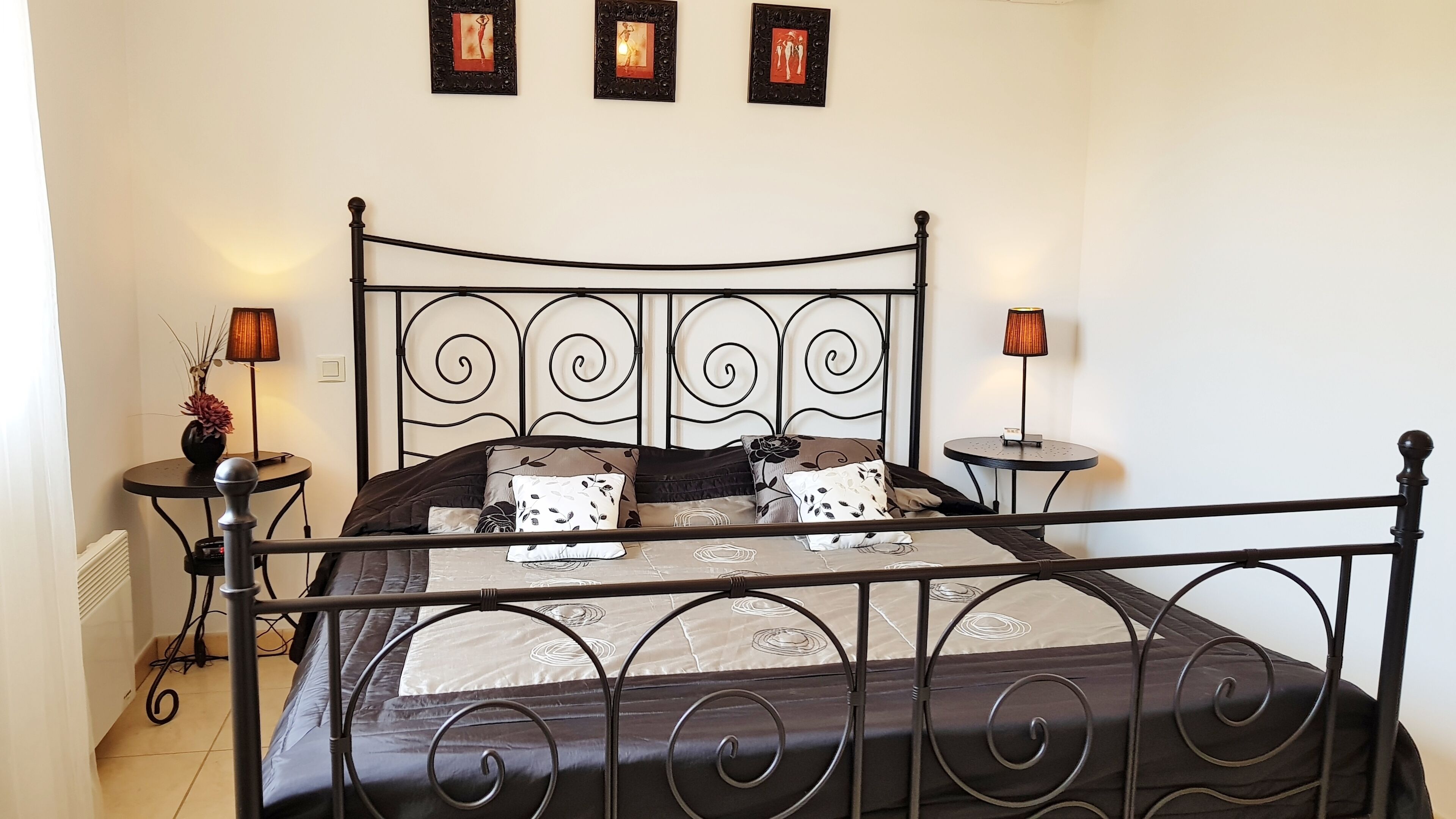 A bedroom with a metal bed frame, a round bedside table with a lamp, and a small table with a lamp. There are three framed pictures on the wall.