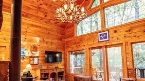 A log cabin interior with a fireplace, wooden furniture, and large windows. - Rushing Creek Cabin, Private Haven, 5 Acres to Explore (Ellijay)