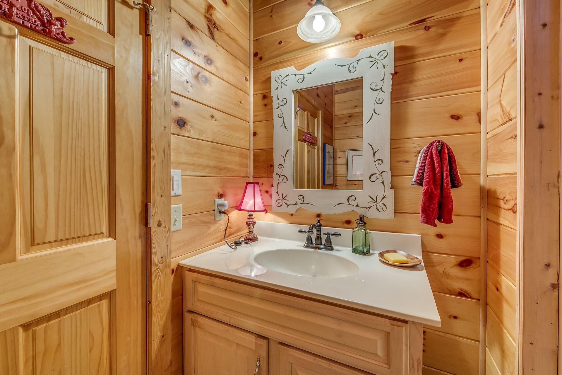 A wooden bathroom with a white sink, a mirror with decorative frame, a pink lamp, and a towel rack with towels.
