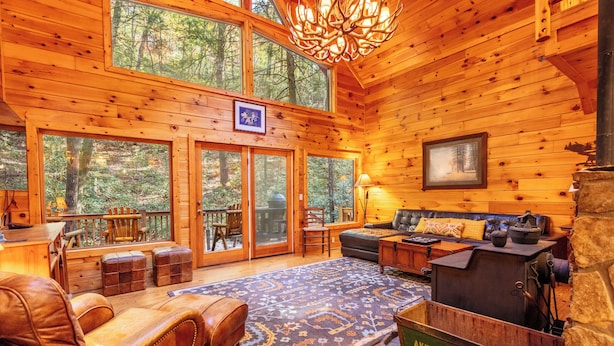 A cozy cabin interior with wooden walls, a fireplace, leather furniture, and large windows overlooking a forest. - Rushing Creek Cabin, Private Haven, 5 Acres to Explore (Ellijay)