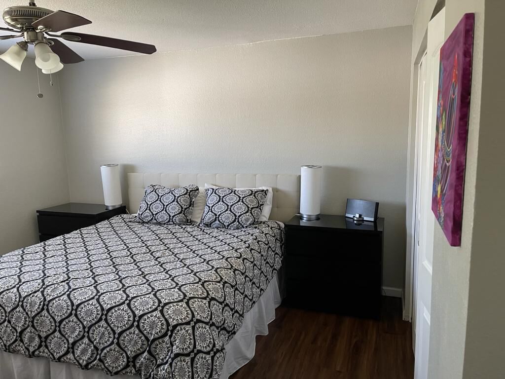 A bedroom with a patterned bedspread, two bedside lamps, a ceiling fan, and a painting on the wall.