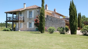 Una casa de piedra con balcón, rodeada por un césped cuidado y altos cipreses.