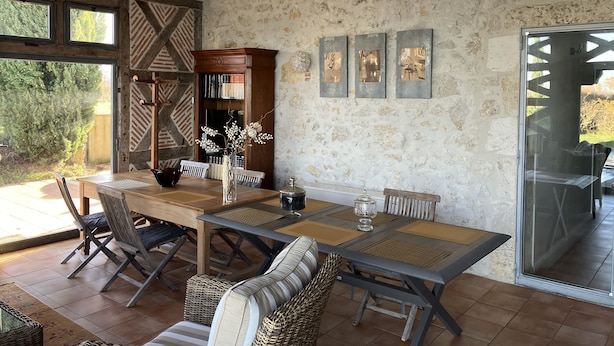 Un comedor con una mesa y sillas de madera, un jarrón con flores y una estantería frente a una pared de piedra.