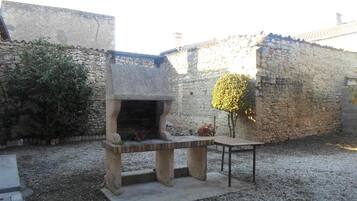 Een buitenbarbecue van natuursteen met een tafel en een bank, omgeven door grind en een stenen muur.