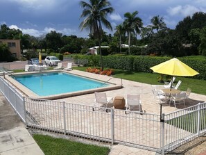 Une piscine rectangulaire entourée d’une clôture blanche, avec des chaises longues blanches et un parasol jaune.